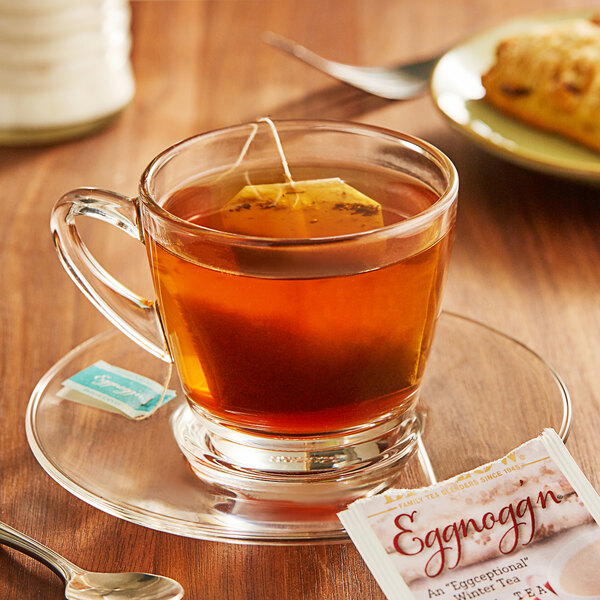 A glass cup of Bigelow Eggnogg'n Tea on a saucer with a tea bag next to a plate of cookies.