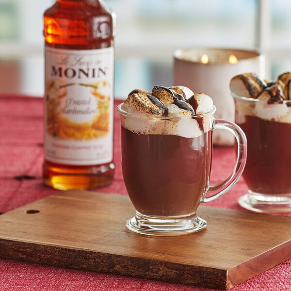 A glass mug of Monin Toasted Marshmallow hot chocolate with a bottle of syrup on the table.