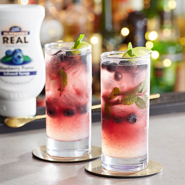 Two glasses of blueberry drinks garnished with mint and blueberries on a table with a bottle of Real Blueberry Puree Infused Syrup.