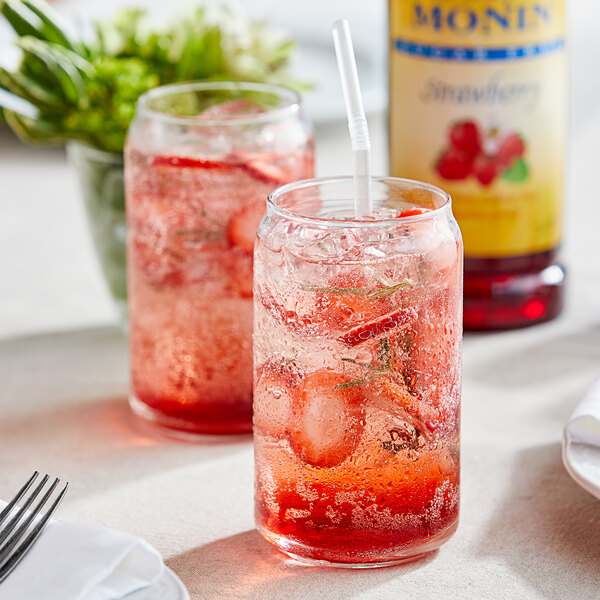 Two glasses of red liquid with straws on a table with a bottle of Monin Sugar Free Strawberry Fruit Syrup.