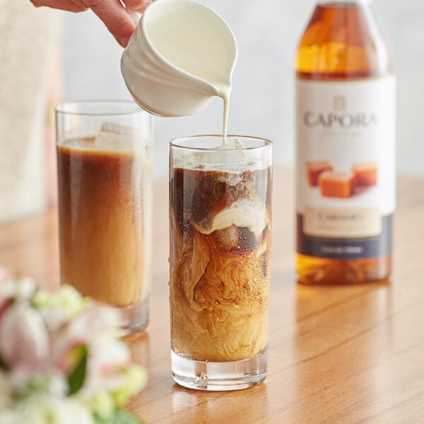 A person pouring Capora caramel flavoring syrup into a glass of brown liquid.