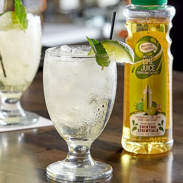 A bottle of Master of Mixes sweetened lime juice on a table with a glass of ice and lime juice.