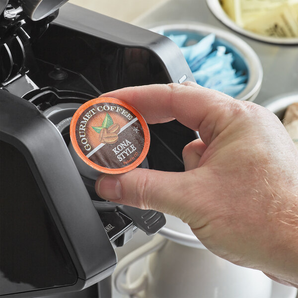 A hand using a device to insert a Caffe de Aroma Kona Style coffee pod into a coffee maker.