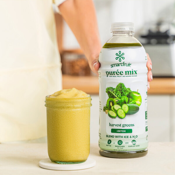 A person's hand holding a bottle of Smartfruit Harvest Greens puree next to a white jar of green smoothie.