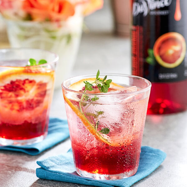 two glasses of red liquid with a slice of orange and a sprig of basil