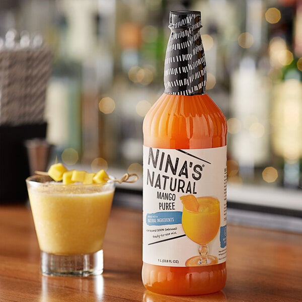 A bottle of Nina's Natural mango puree on a table with a glass of yellow liquid and fruit pieces.