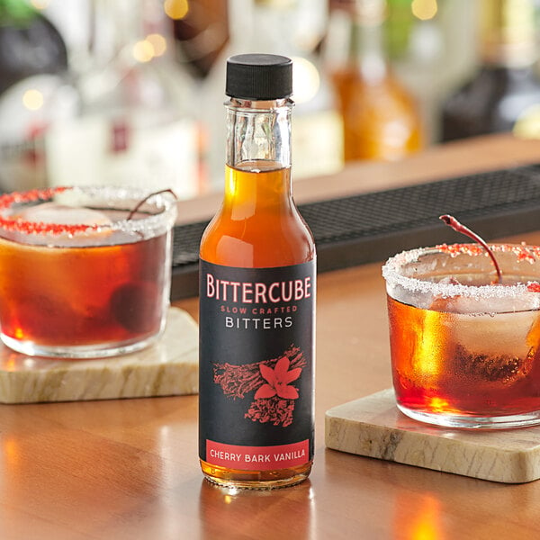 A bottle of Bittercube Cherry Bark Vanilla Bitters on a table with glasses of liquid.