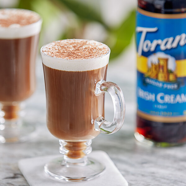 A glass mug of coffee with brown liquid and foam with a bottle of Torani Sugar-Free Irish Cream flavoring syrup.