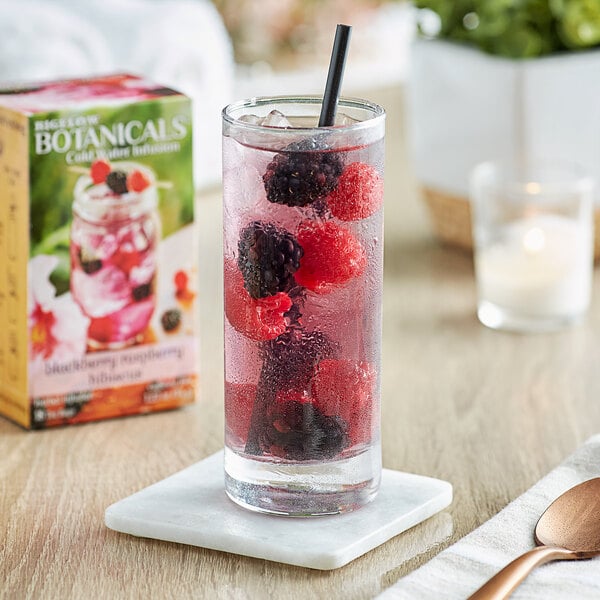 A glass of Bigelow Botanicals Blackberry Raspberry Hibiscus Cold Water Infusion Tea with berries and ice next to a box of the tea.