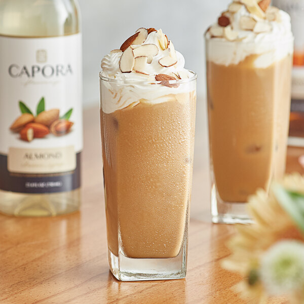 Two glasses of iced coffee with Capora Almond Flavoring Syrup and vanilla next to a bottle of Capora Almond Flavoring Syrup.