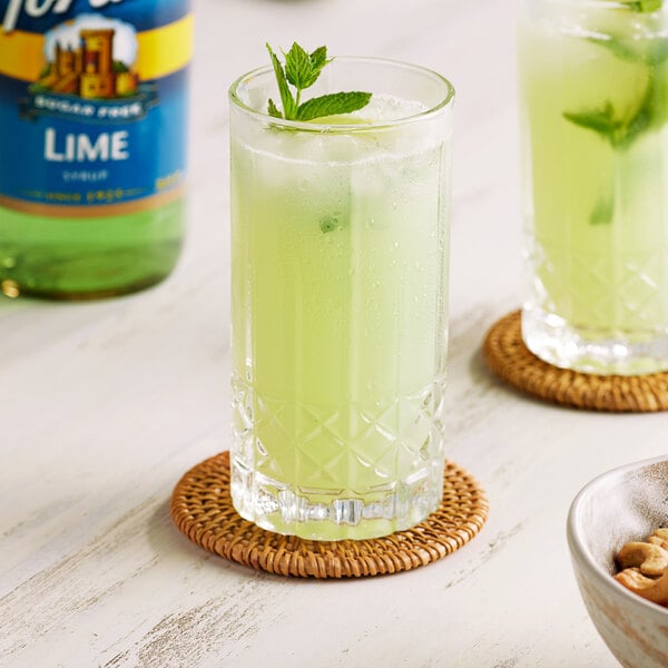 A glass of green liquid with mint leaves and a straw on a table with a Torani Sugar-Free Lime Fruit Syrup bottle.