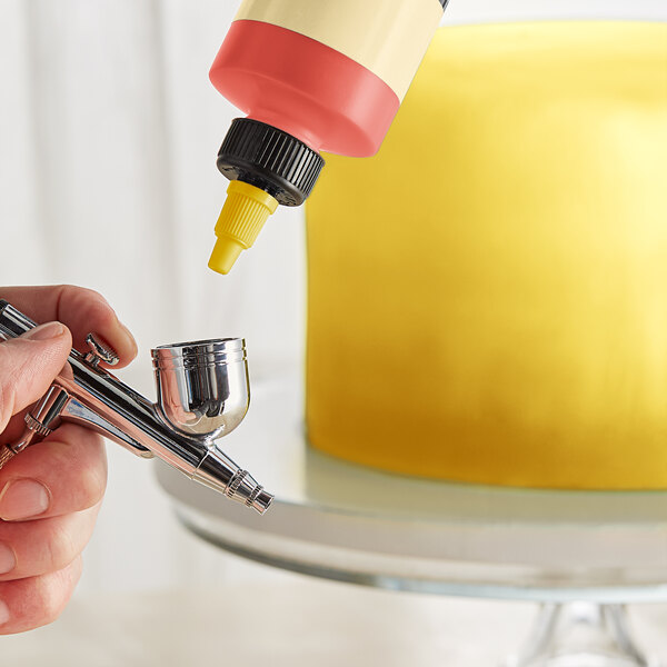 A hand using a silver Chefmaster airbrush to spray yellow food coloring over a cake.