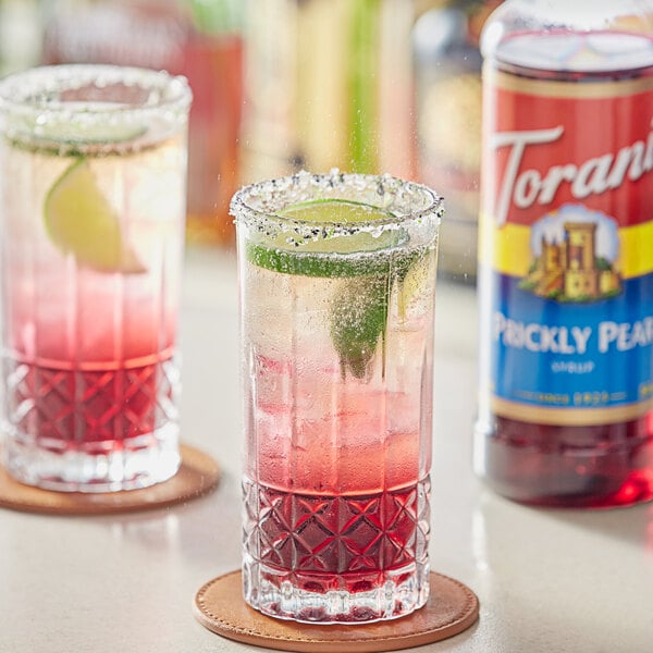 Two glasses of a pink drink with a green leaf and lime next to a bottle of Torani Prickly Pear Flavoring Syrup.