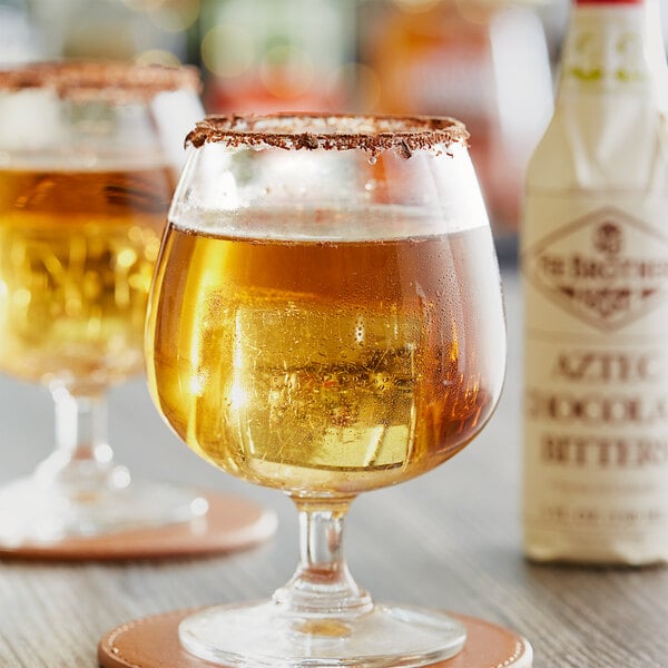 Two glasses of brown liquid on a table in a bar with a bottle of Fee Brothers Aztec Chocolate Bitters.