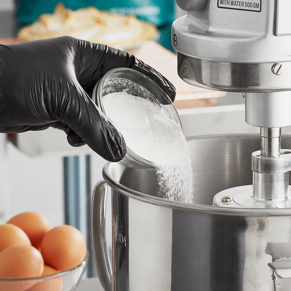 A person in black gloves pouring Judee's meringue powder into a metal container on a professional kitchen counter.