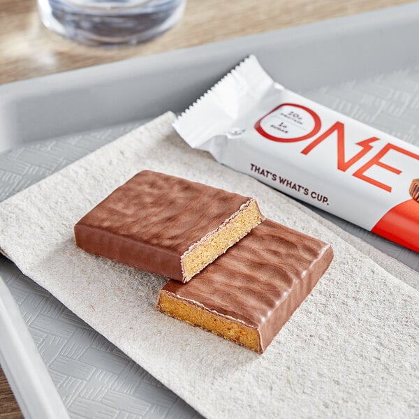 A tray with a chocolate bar on it next to a white package of ONE Peanut Butter Cup Protein Bars.