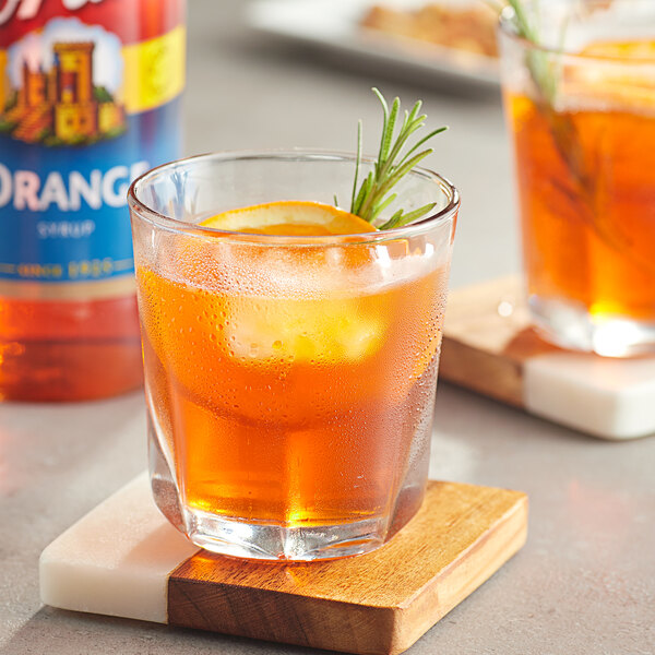 A glass of orange liquid with a rosemary sprig on a wooden coaster.