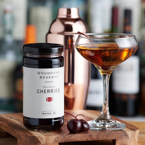 A jar of Woodford Reserve Bourbon Cherries on a wooden tray with a cocktail glass filled with a cherry drink.