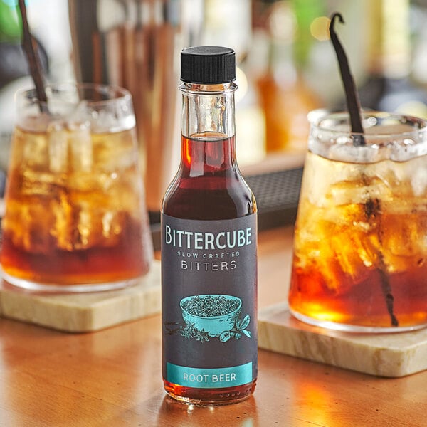 A bottle of Bittercube Root Beer Bitters on a table next to two glasses of brown liquid.