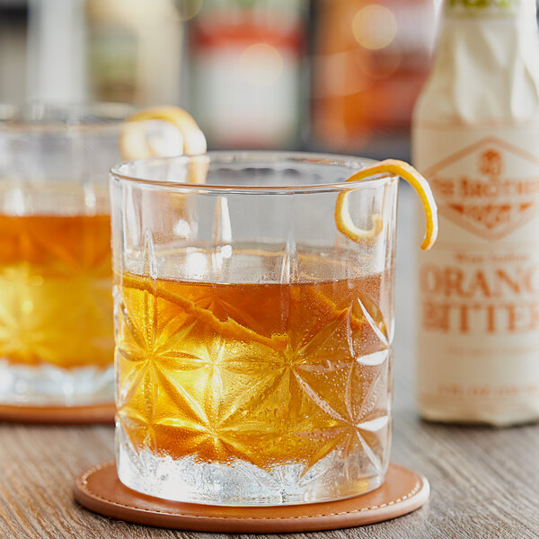 Two glasses of whiskey with orange slices next to a bottle of Fee Brothers Orange Bitters.