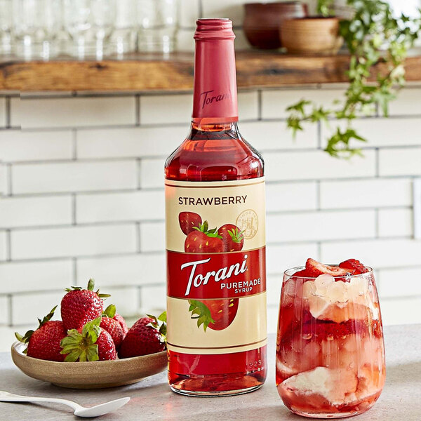 A bottle of Torani strawberry syrup next to a glass of ice and strawberries.