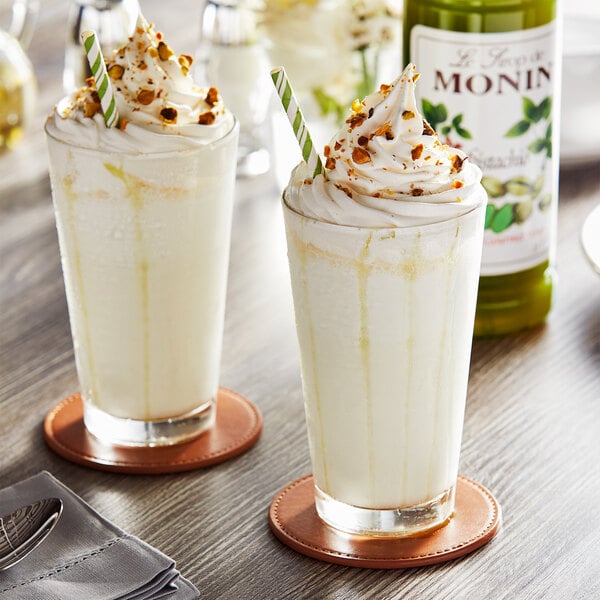 Two glasses of milkshakes with whipped cream and green and white striped straws on a table with a Monin Pistachio Syrup bottle.