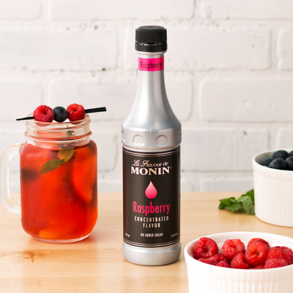 A Monin Raspberry Concentrated Flavor bottle on a table next to a bowl of raspberries.