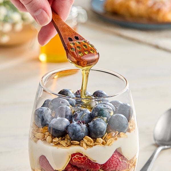 A wooden spoon drizzling Hometown Provisions all-natural honey over a glass of yogurt with blueberries.