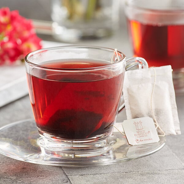 A glass cup of Wild Hibiscus Heart-Tee herbal tea with a tea bag on a saucer.