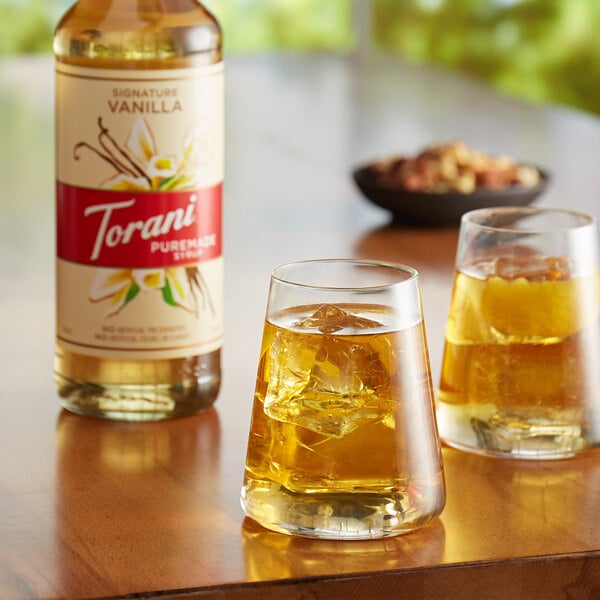 A glass of Torani Puremade Signature Vanilla flavoring syrup and ice next to a bottle of the syrup on a table with another glass of liquid.