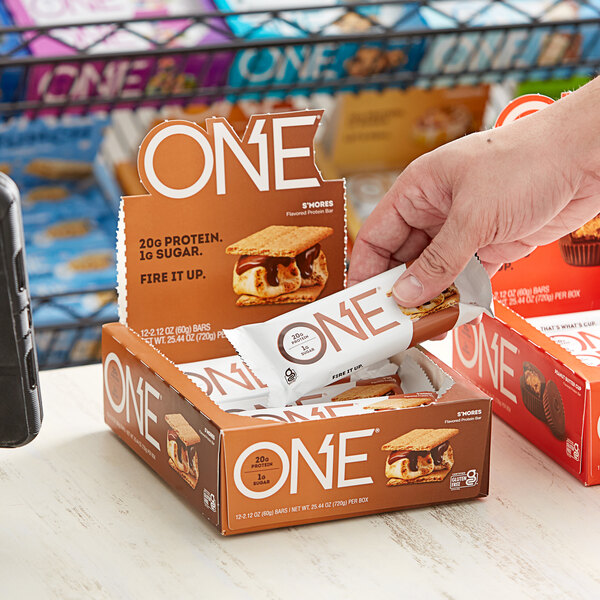 A hand holding a box of ONE S'mores Protein Bars on a table.