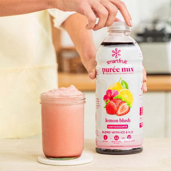 A person holding a bottle of Smartfruit Lemon Blush puree next to a glass of pink liquid.