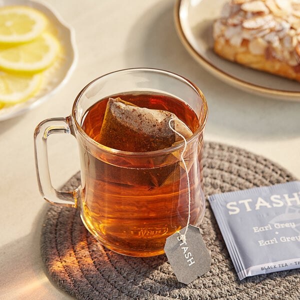 A glass mug of Stash Earl Grey tea with a tea bag on the table next to a plate of lemon slices.