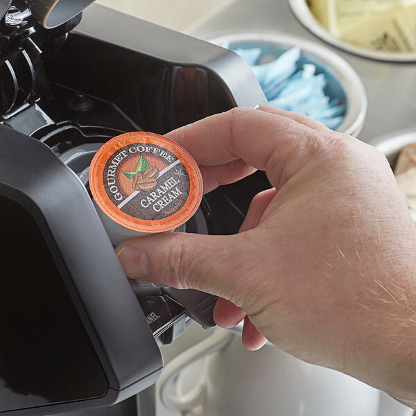 A hand placing a Caffe de Aroma Caramel Cream coffee capsule into a coffee maker.