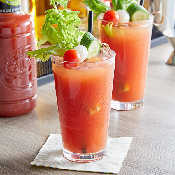 Two glasses of Finest Call Zesty Bloody Mary Mix with vegetables and a straw next to a red container of the mix.