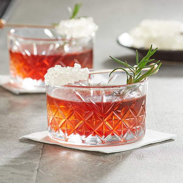 A glass of red drink with a Roses Dryden and Palmer white swizzle stick and a rosemary sprig.