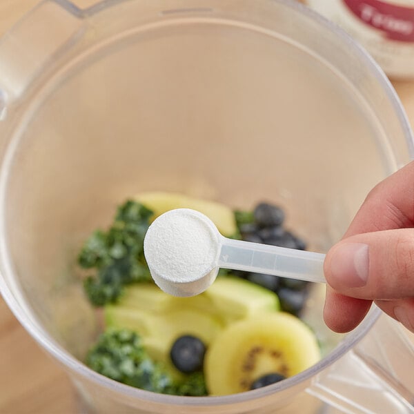 A hand using a measuring spoon to add white supplement powder to a blender.