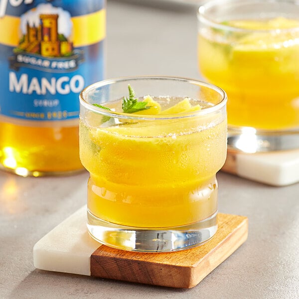 A glass of yellow liquid with a leafy green leaf on top next to a bottle of Torani Sugar-Free Mango Fruit Syrup.