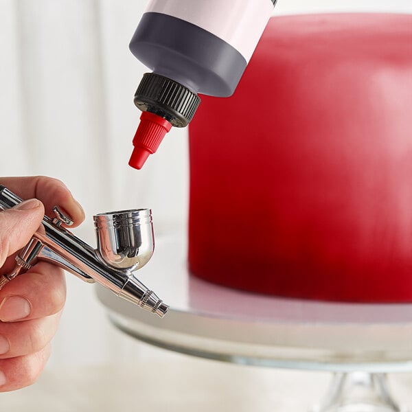 A hand using a silver Chefmaster airbrush to spray red food coloring over a cake.