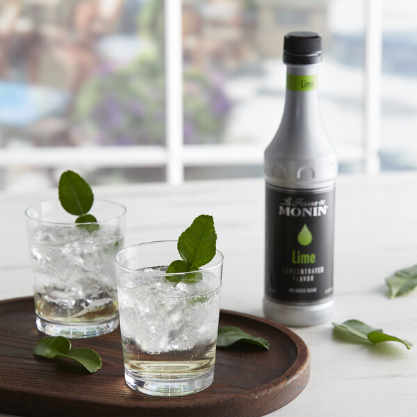 A wooden tray with two glasses of ice and Monin Lime Concentrated Flavor with leaves.