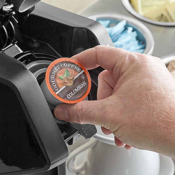 A hand holding a round orange and black Caffe de Aroma Colombian Supreme coffee capsule.