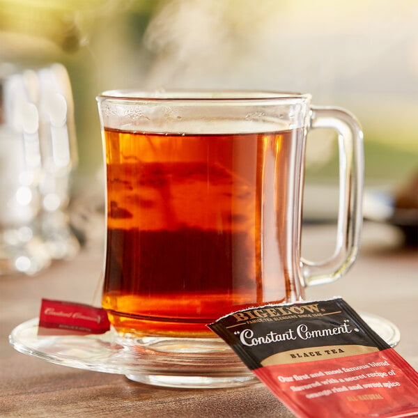 A glass mug of Bigelow Constant Comment tea on a saucer with a tea bag on the table next to a packet of tea.