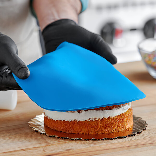 A person wearing black gloves and using a blue spatula to cut a cake covered in blue Satin Ice fondant.