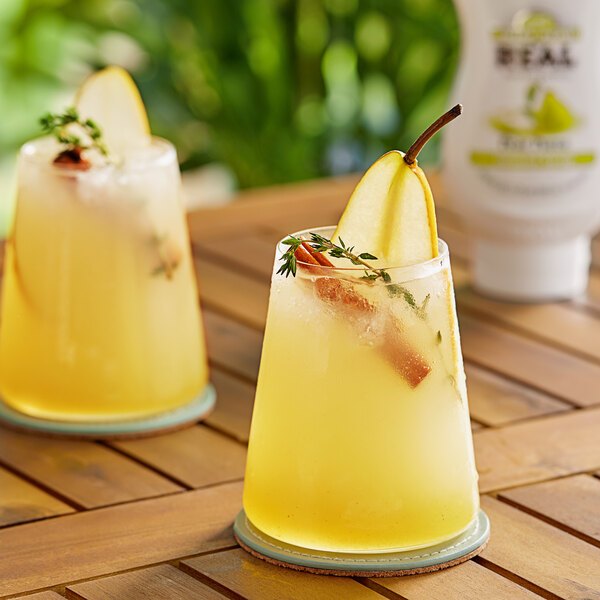 A pair of glasses of yellow liquid with pear slices on a table.