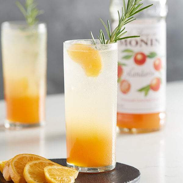 A close-up of a glass of Monin mandarin syrup flavored orange juice with a rosemary sprig on top, garnished with a slice of orange.