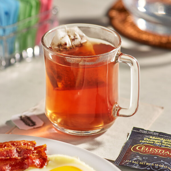 A glass mug of Celestial Seasonings Mandarin Orange Spice Tea with a tea bag on a plate of breakfast food.