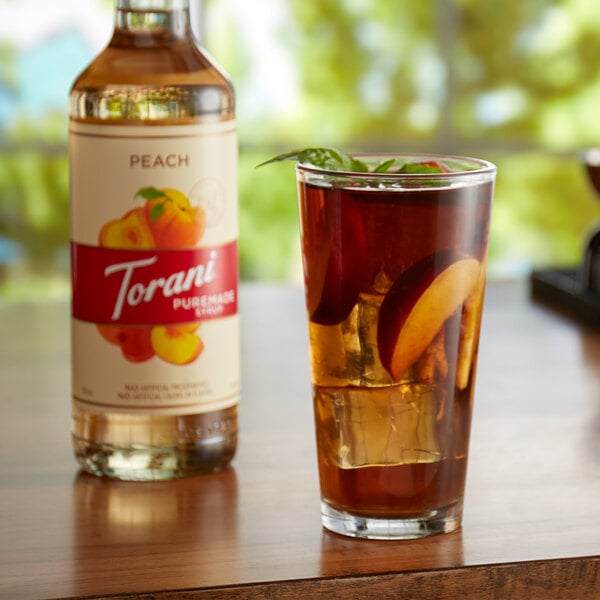 A glass of peach tea with ice and fruit next to a Torani Puremade Peach Flavoring Syrup bottle.