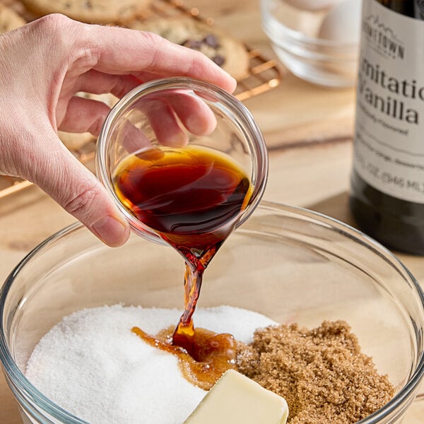 A hand pouring brown liquid from a Hometown Provisions Imitation Vanilla bottle into a glass bowl of brown sugar.