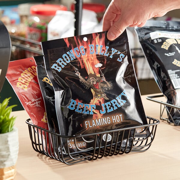 A hand holding a black bag of Bronco Billy's Flaming Hot Beef Jerky with a cartoon character label.