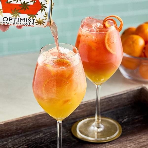 A person pouring orange and yellow Optimist Botanicals Non-Alcoholic Cali Spritz into a glass.